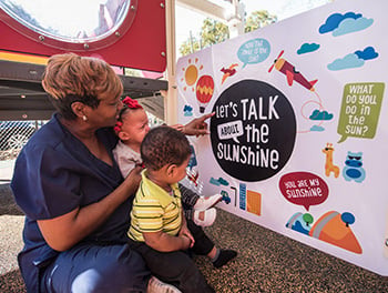 Two toddlers and an adult explore a Talking is Teaching panel, an ideal inclusive playground example.
