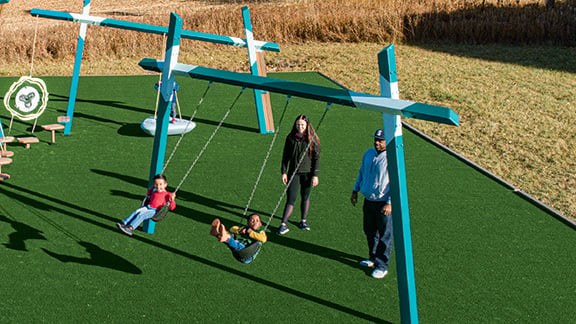Two children swinging on a Forma® swing frame while a parent pushes them.