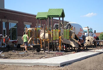 Workers shown installing a PlayShaper play system at a school. Playground has green roofs to provide shade, a crawl tunnel and poly slide. 