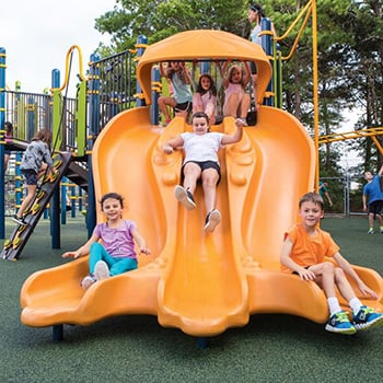 Three children slide down the Cloudburst playground equipment slide, which is a three-bed polyethylene colorful multi-user slide