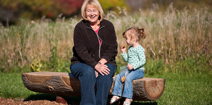 A woman with glasses sits next to a young girl eating a snack on a log bench.