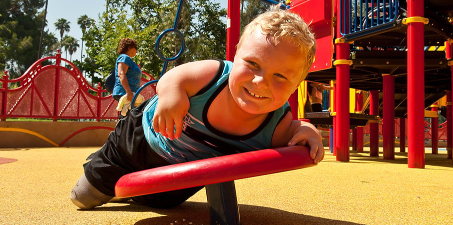 Young boy smiles at the camera as he lays across a pod climber at an inclusive playground.