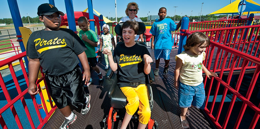 Boy smiling in wheelchair while wheeling down a inclusive PlayBooster playground decking with a group of children.