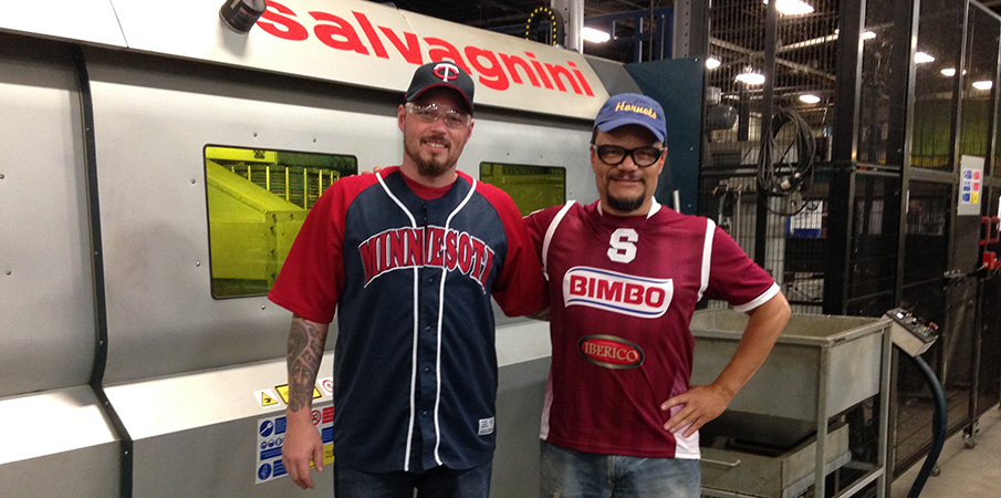 Two Landscape Structures employees smile for the camera while standing in front of a large manufacturing machine wearing team jerseys for Team Spirit Day.